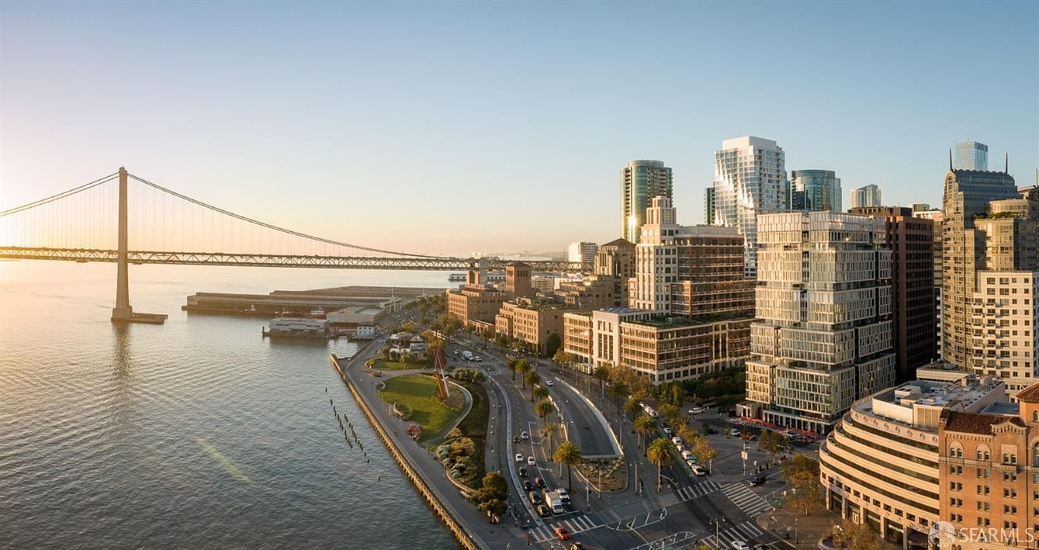 This aerial view showcases a stunning cityscape with a prominent suspension bridge extending over the water. Modern buildings with unique architectural designs dominate the skyline, complemented by lush green spaces along the waterfront. The image captures a vibrant urban environment with a blend of natural and man-made elements.