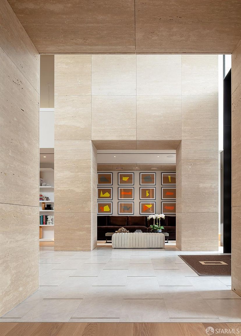 This interior shot showcases a grand hallway with travertine walls and flooring, creating a luxurious and spacious feel. The hallway leads to a seating area adorned with framed artwork and a plush bench, suggesting a sophisticated and welcoming atmosphere. The architectural design emphasizes clean lines and a neutral color palette, enhancing the sense of elegance and tranquility.
