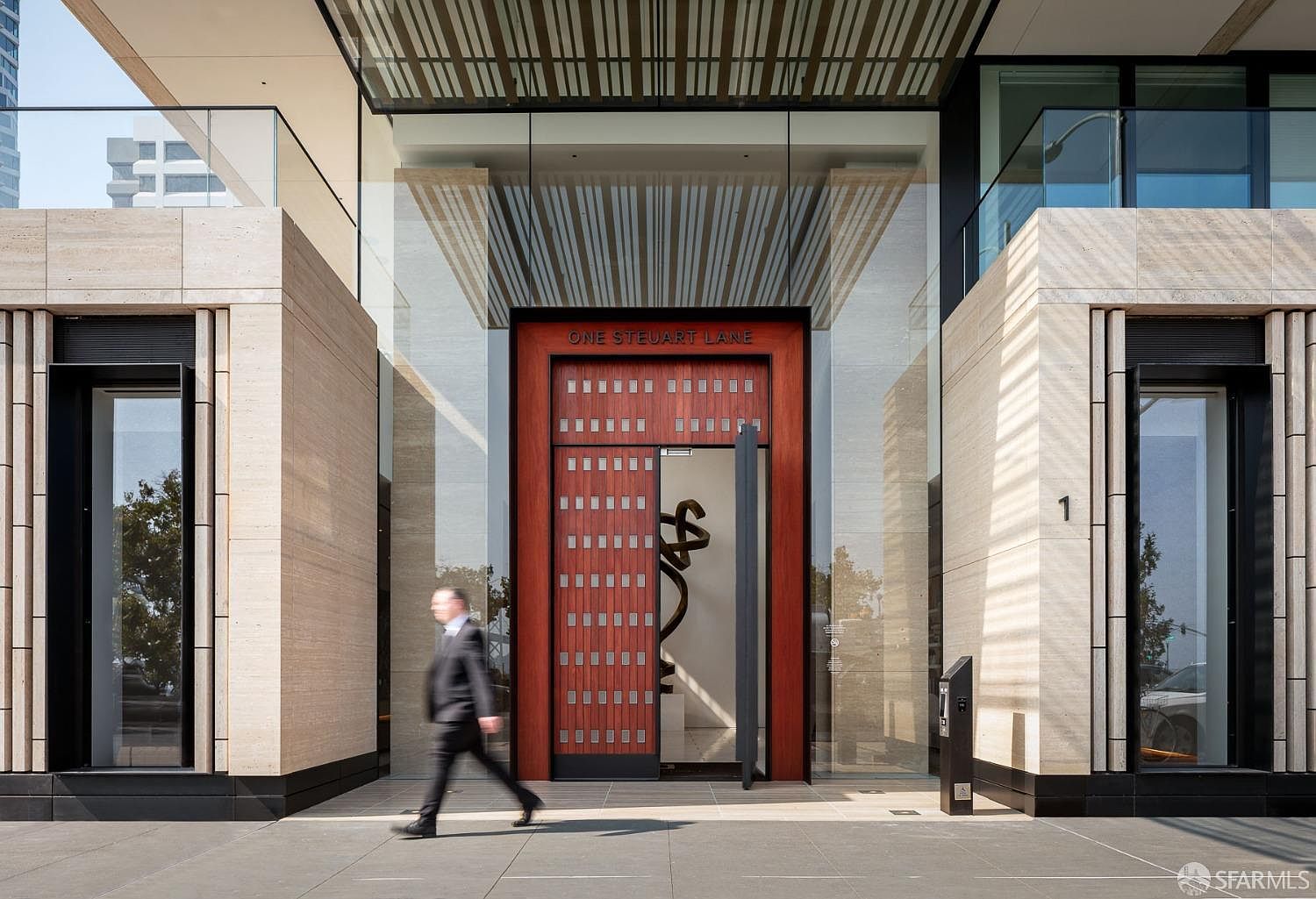 The image showcases the grand entryway of a modern building, featuring a large wooden door with metallic accents and the address "ONE STEUART LANE" inscribed above. The entrance is flanked by glass panels and stone-clad walls with vertical detailing, creating a sophisticated and inviting aesthetic. A blurred figure walking by adds a sense of scale and activity to the scene.
