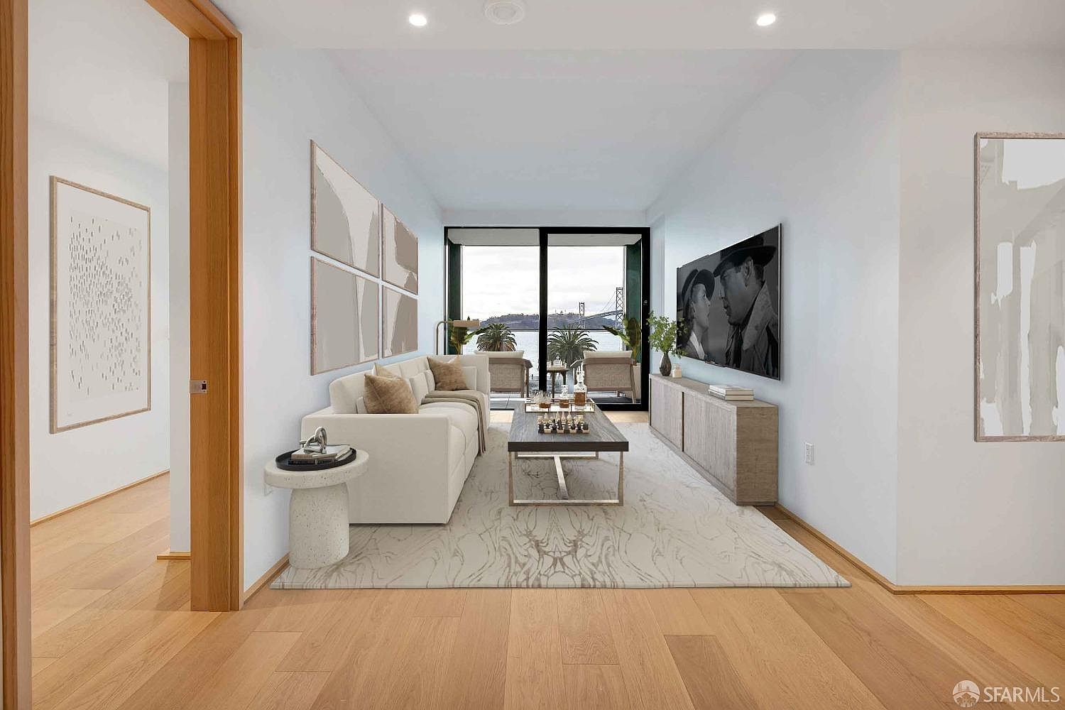 This is a bright and modern living room featuring a neutral color palette with hardwood floors and white walls. The room is furnished with a white sectional sofa, a contemporary coffee table, and a media console with a large television. Large windows offer a view of the water and bridge, enhancing the room's appeal and creating a serene atmosphere.