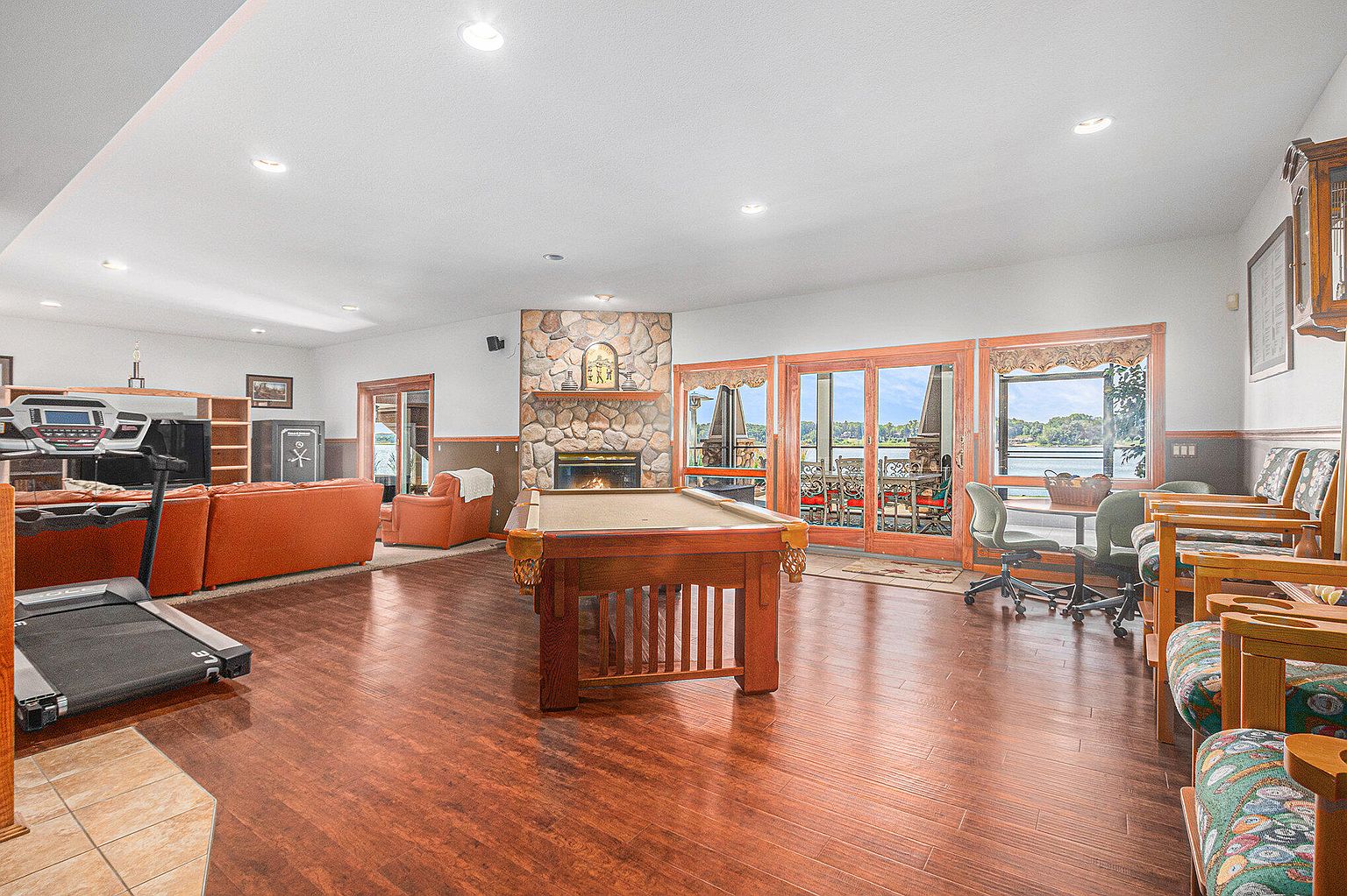 This is a spacious basement featuring a pool table, a stone fireplace, and a seating area with an orange sofa. The room also includes a treadmill, a dining table with chairs near large windows offering a view of the lake, and hardwood floors throughout. The overall impression is a comfortable and versatile living space.