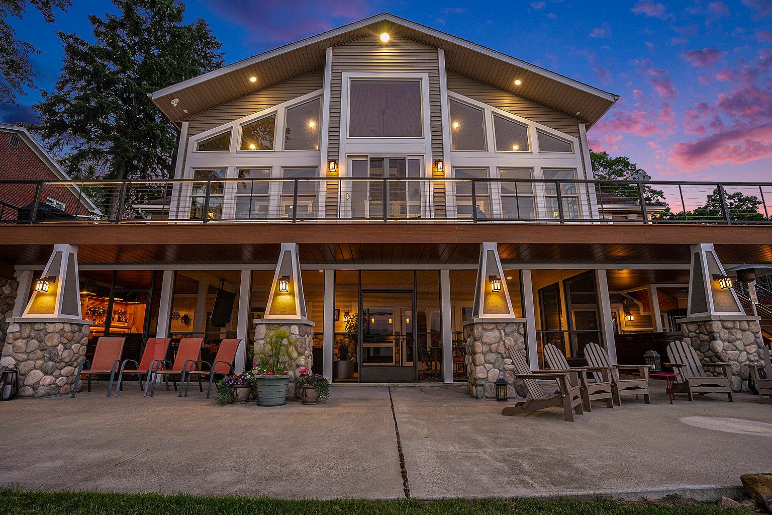 This is a rear view of a two-story house featuring a large deck on the second floor with a metal railing. The lower level has a patio with stone pillars and outdoor seating. The house has many large windows, and the exterior is a mix of siding and stone.