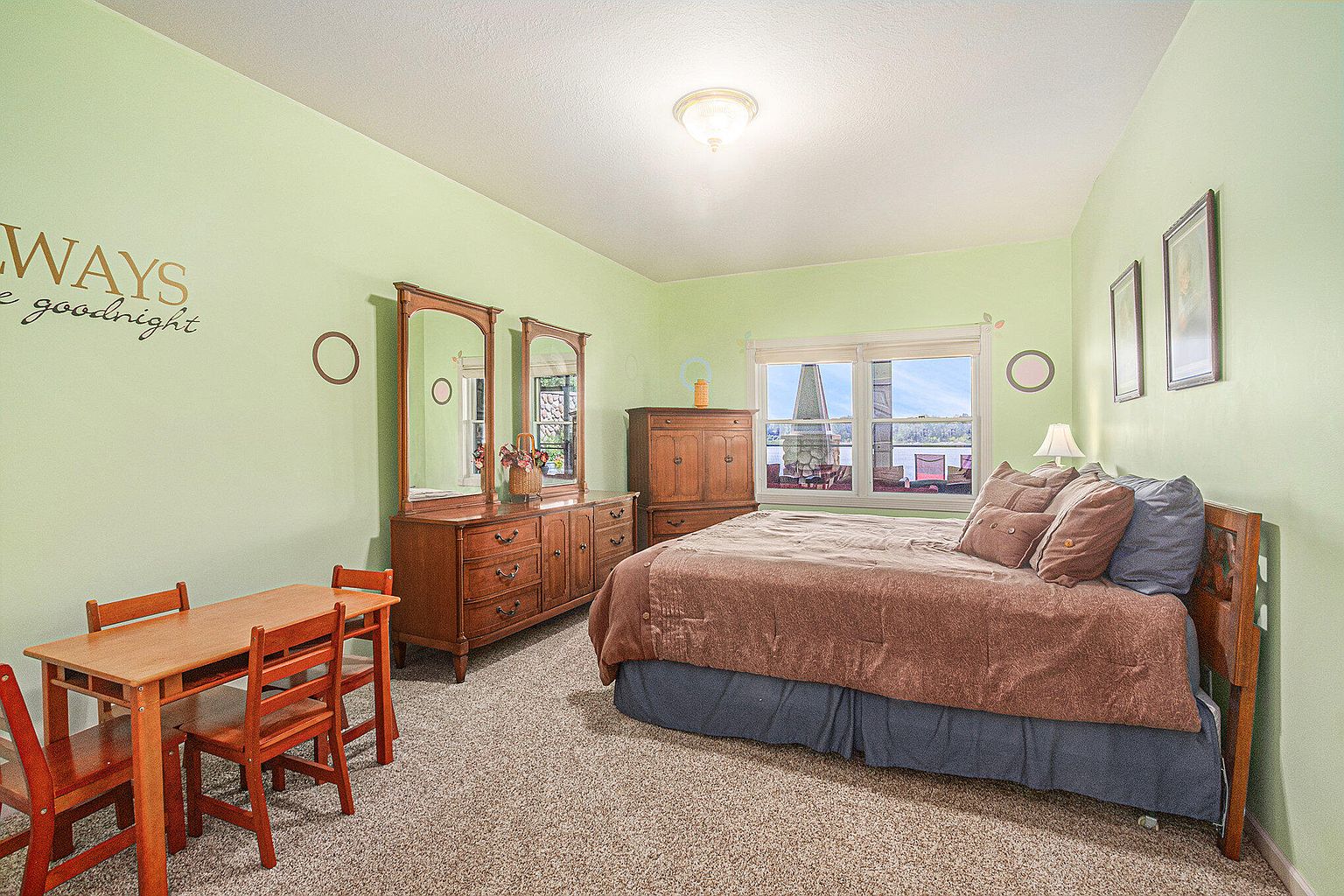 This is a cozy bedroom featuring light green walls, a carpeted floor, and a wooden bed with brown bedding and pillows. The room includes a wooden dresser with mirrors, a small wooden table with chairs, and framed artwork on the walls. A window provides natural light and a view of the outdoors, creating a comfortable and inviting space.