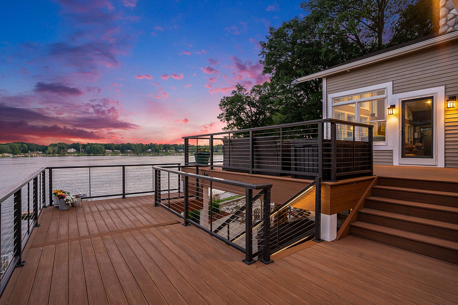 This image showcases a beautifully designed outdoor deck and balcony area, perfect for lakeside living. The deck features modern cable railings, warm-toned wood flooring, and steps leading to a lower level. A portion of the deck is elevated, creating a separate seating area, and the adjacent house has a well-lit entrance and large windows, all set against a stunning sunset backdrop.