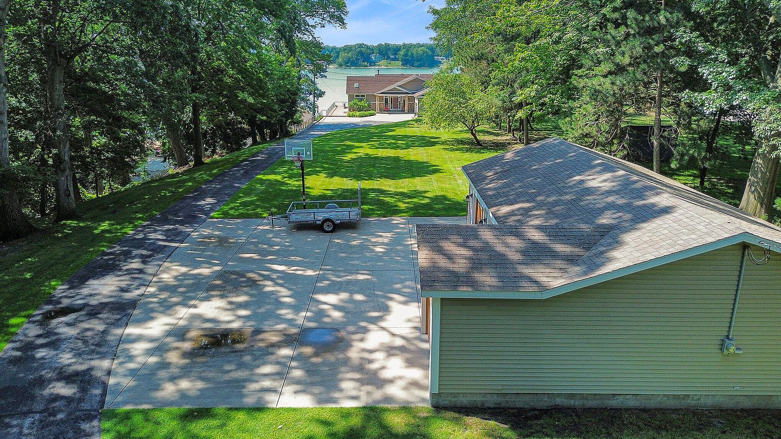 This aerial shot showcases a well-maintained property featuring a spacious concrete driveway, a basketball hoop, and a trailer. The house has a light green exterior with a shingled roof, and the surrounding landscape includes lush green lawns and mature trees. In the background, a glimpse of a lake and another house adds to the property's appeal.
