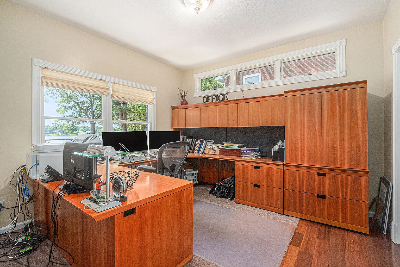 This is an interior shot of a home office featuring a large wooden desk with computer monitors and office supplies. The room includes built-in wooden cabinetry and shelving, providing ample storage. A window offers natural light and a view of the outdoors, creating a comfortable and functional workspace.