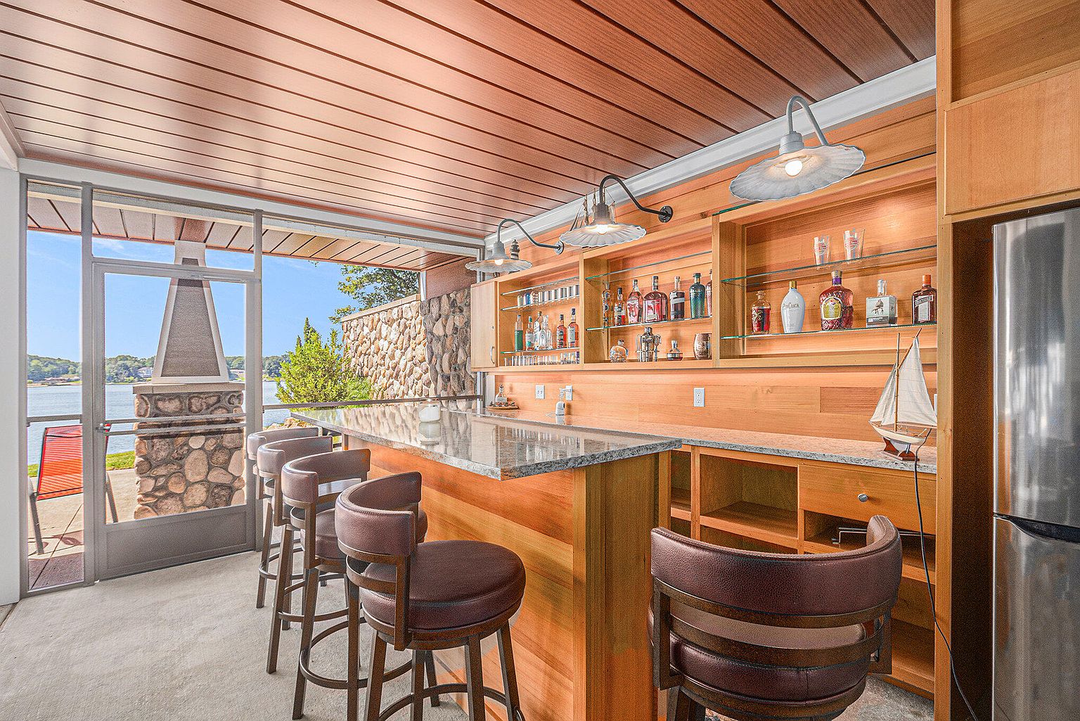 This sunroom features a stylish bar area with wood cabinetry, granite countertops, and open shelving displaying various bottles. Bar stools line the counter, offering seating with a view of the lake through the open doorway. The room has a warm, inviting atmosphere with natural light and a relaxed, upscale feel.