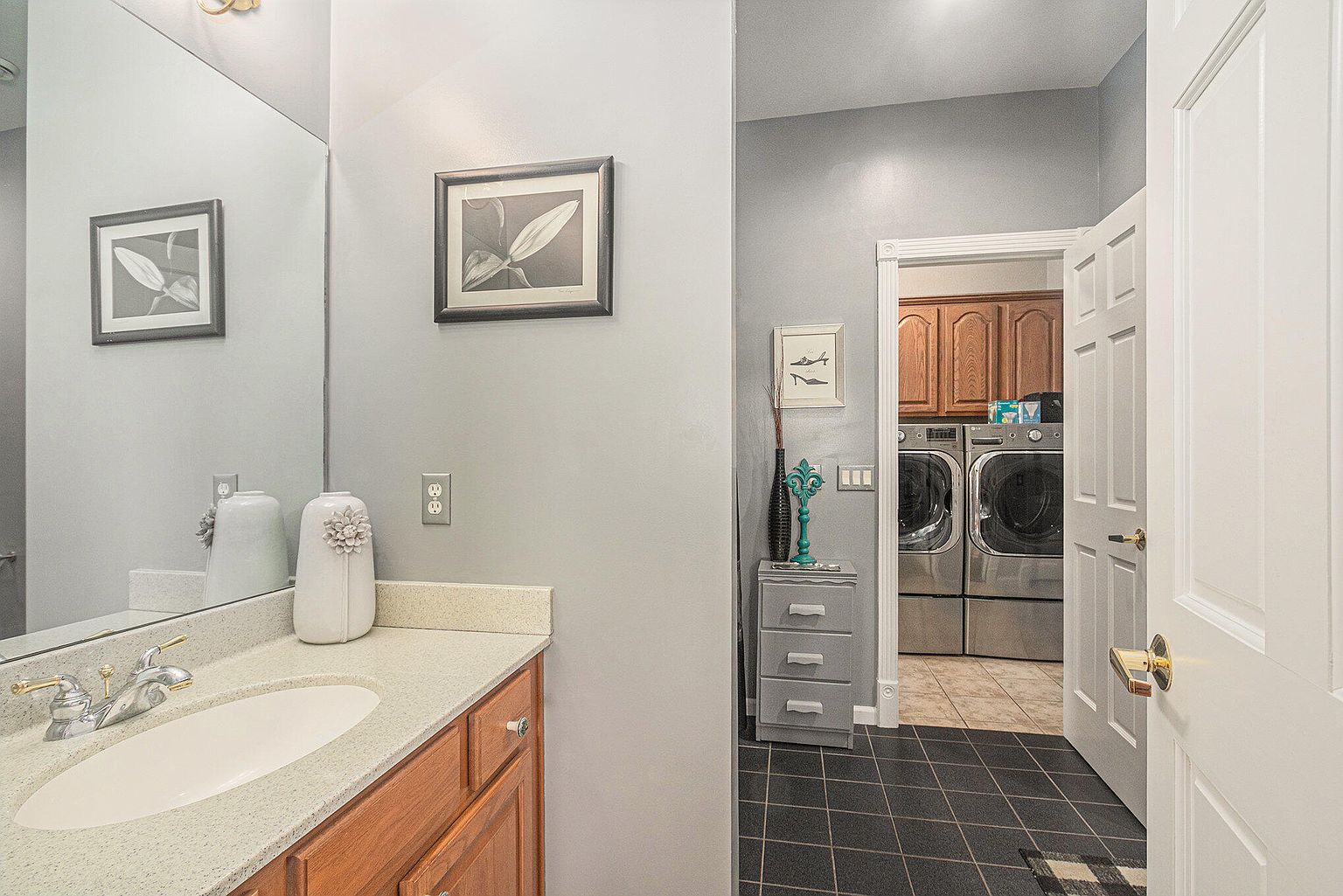 This is a well-lit guest bathroom that leads into a laundry room. The bathroom features a wooden vanity with a light countertop, a sink, and a large mirror. The laundry room is visible through the doorway and contains a washer and dryer set with wooden cabinets above.