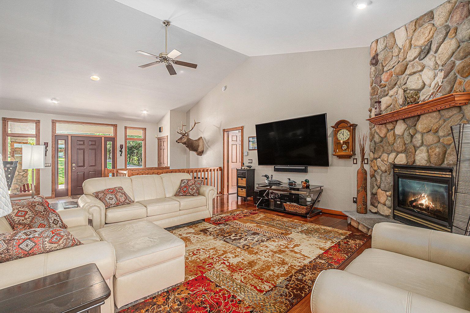 This is a warm and inviting living room featuring a stone fireplace, hardwood floors, and comfortable white leather furniture. A large television is mounted above a media console, and a decorative rug adds a touch of color. The room has high ceilings and a ceiling fan, creating an open and airy feel.