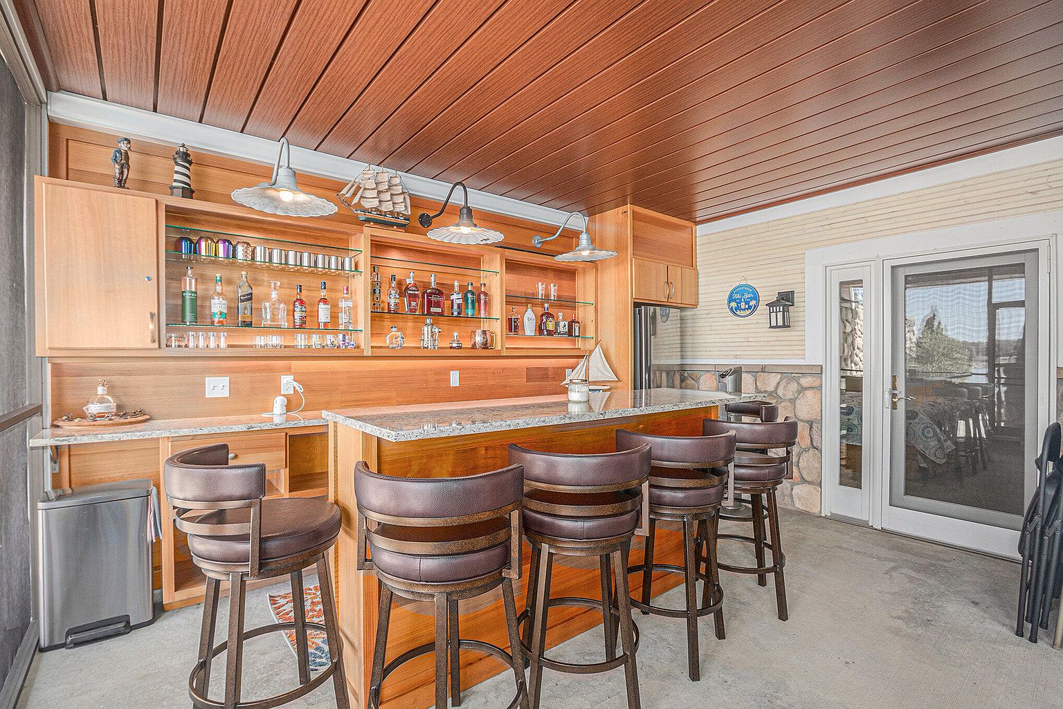 This image showcases an indoor bar area within a sunroom or conservatory. The bar features wooden cabinetry, glass shelves displaying liquor bottles, and a granite countertop with bar stools. The space is well-lit with overhead lighting and natural light from the adjacent outdoor area, creating a relaxed and inviting atmosphere.