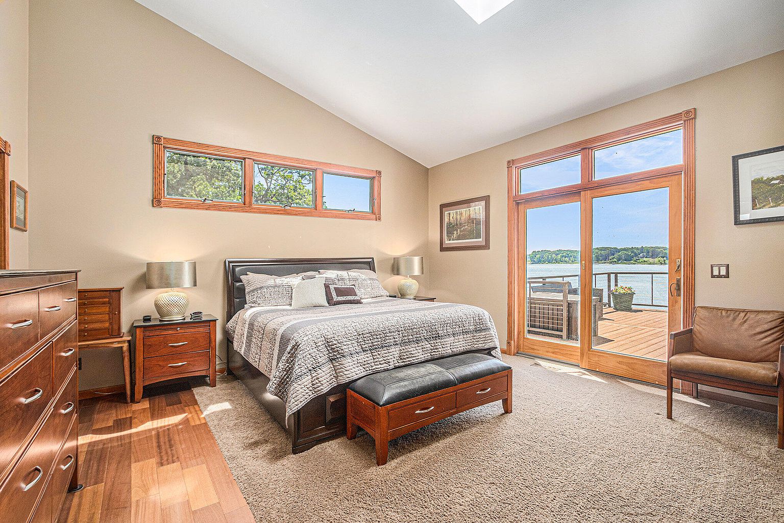 This is a primary bedroom featuring a king-size bed with a patterned comforter and several pillows. The room has a neutral color palette with wood accents, including a window frame and a door leading to a deck. A leather bench sits at the foot of the bed, and there is a nightstand on either side of the bed with lamps. The room has a cozy and inviting atmosphere with a view of the lake.