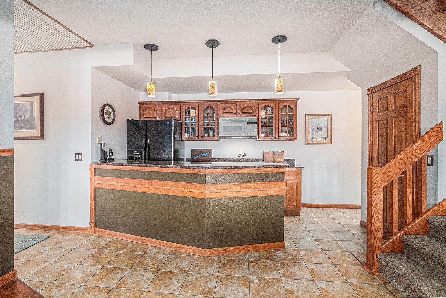 This interior shot showcases a basement area featuring a wet bar with wooden cabinetry, a black refrigerator, and pendant lighting. The flooring is tiled, and a staircase with wooden railings is visible on the right. The overall impression is a cozy and functional entertainment space.