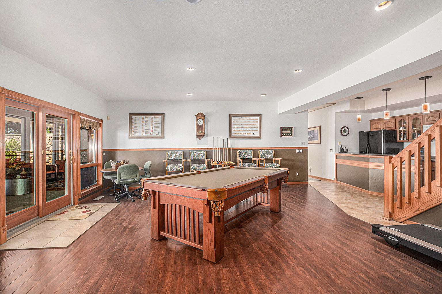 This is a spacious basement featuring a pool table as the focal point, surrounded by a seating area and a partial kitchen. The room has a warm and inviting feel with dark wood flooring and a combination of white and brown wall colors. Natural light enters through a sliding glass door, enhancing the overall ambiance of the space.