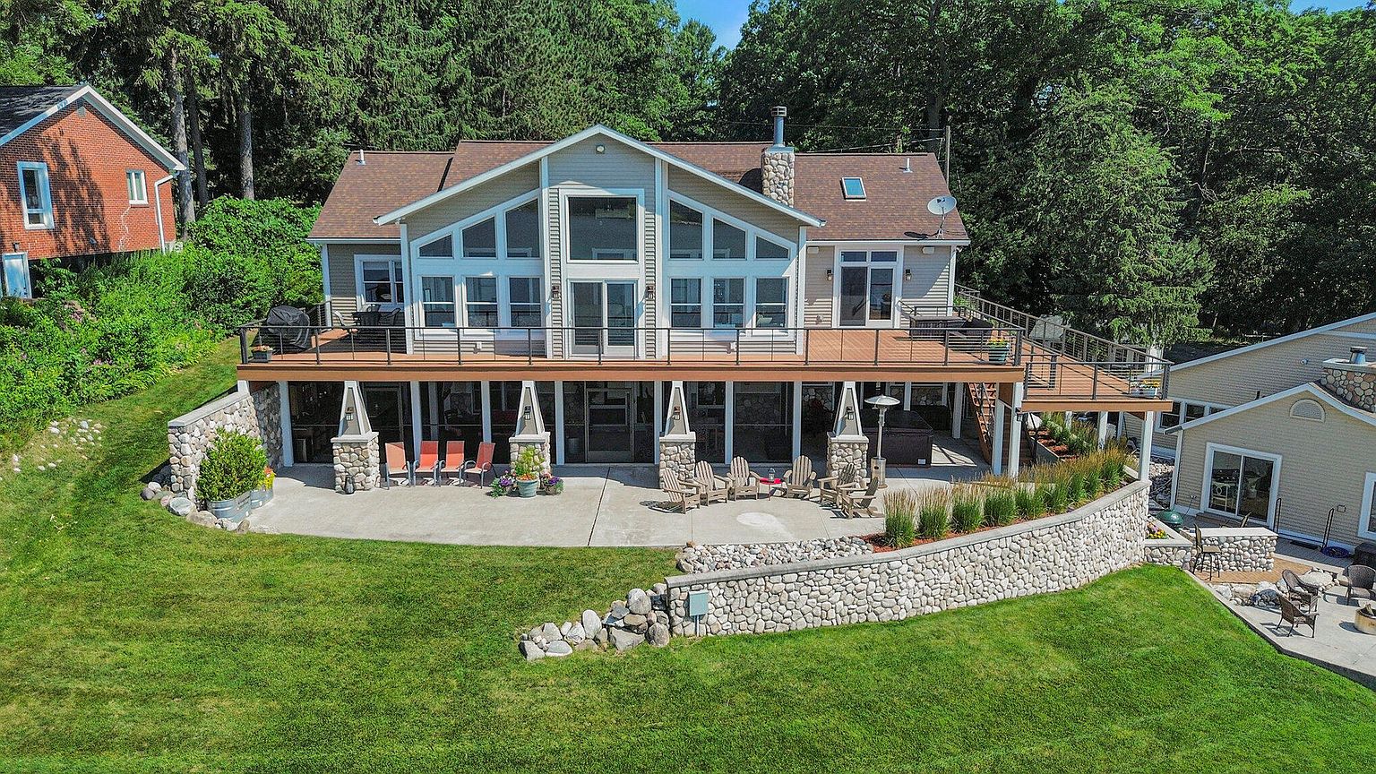 This is an exterior rear view of a two-story house with a large deck and patio area. The house features a combination of siding and stone accents, with large windows providing ample natural light. The backyard includes a well-maintained lawn and stone retaining walls, creating an inviting outdoor living space.