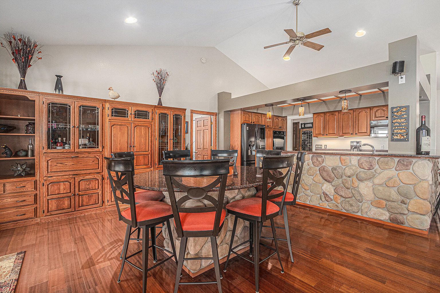 This interior shot showcases a dining area adjacent to a kitchen, featuring a round table with stone base and seating for six. The room is characterized by warm wood flooring and cabinetry, with a stone-clad kitchen counter visible in the background. The space exudes a cozy and inviting atmosphere, perfect for family meals and entertaining.