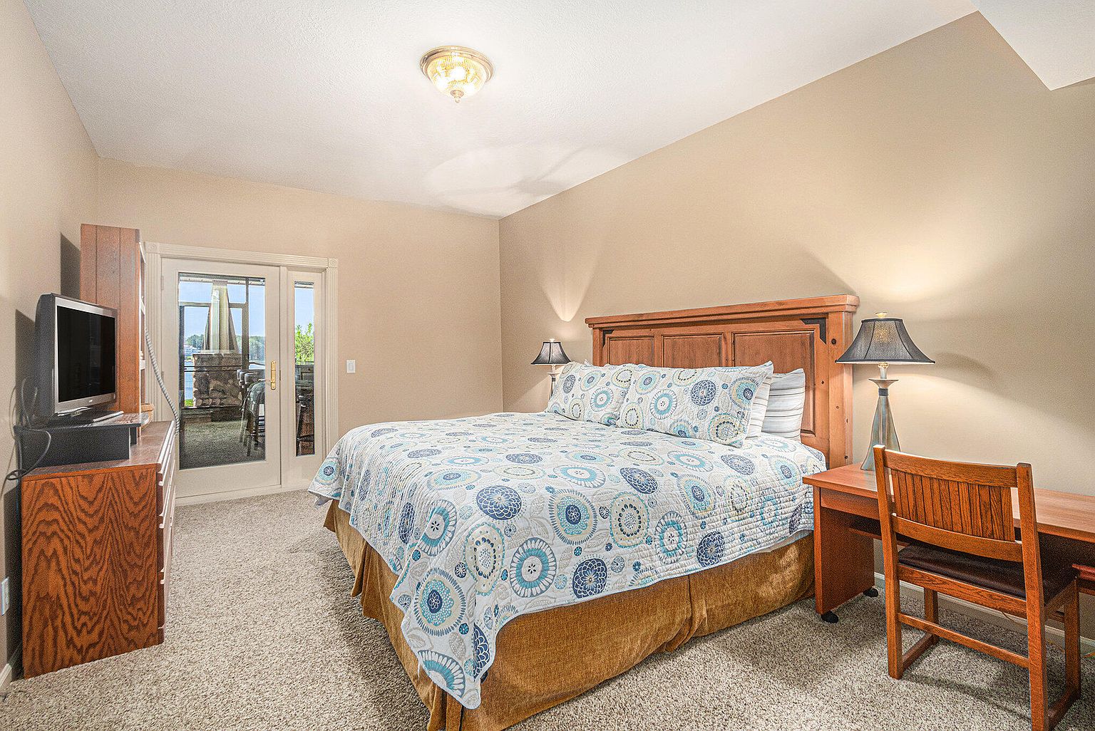 This is a primary bedroom featuring a wooden bed with a patterned blue and white quilt, complemented by matching pillows. A wooden desk and chair sit to the side, illuminated by a lamp, while a TV and dresser are positioned near a sliding glass door that leads to an outdoor patio. The room has a warm, inviting feel with neutral-toned walls and carpet.