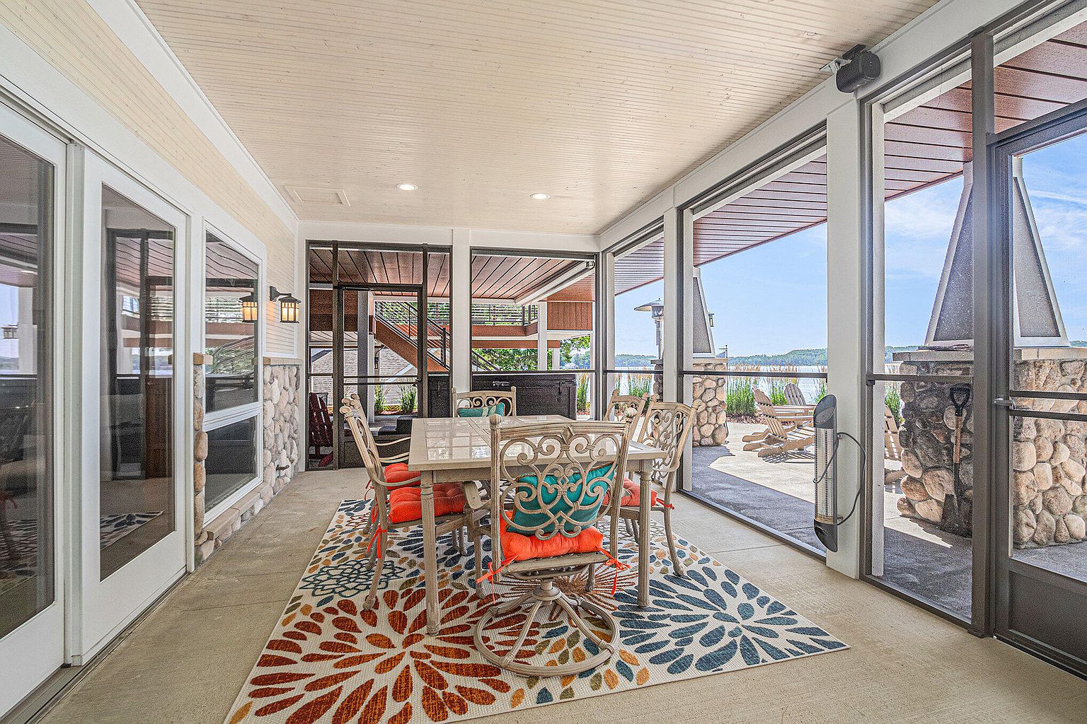 This sunroom features a dining area with a table and chairs set on a colorful floral rug. The room is enclosed with large windows and screens, offering an open view to the outdoor patio and lake. The ceiling is paneled, and the space is well-lit, creating a bright and inviting atmosphere.