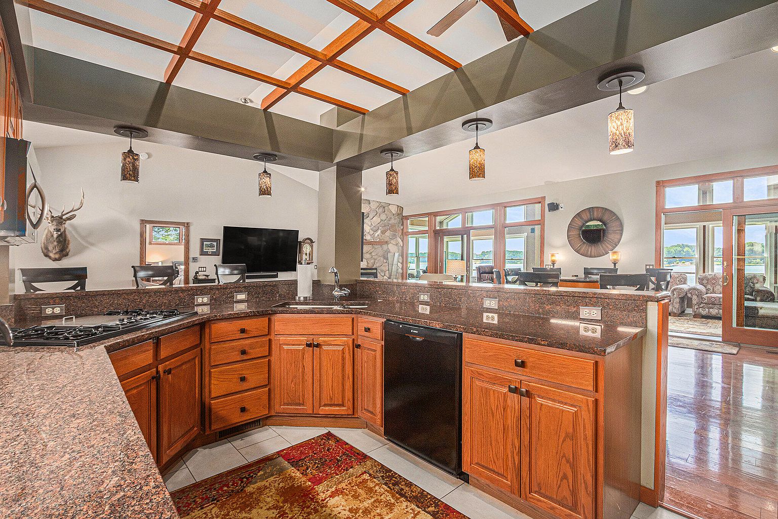 This is a well-appointed kitchen with warm wood cabinetry, granite countertops, and modern appliances. The kitchen features a unique ceiling design with wooden beams and pendant lighting. The space opens to a living area with views of the outdoors, creating an inviting and connected atmosphere.
