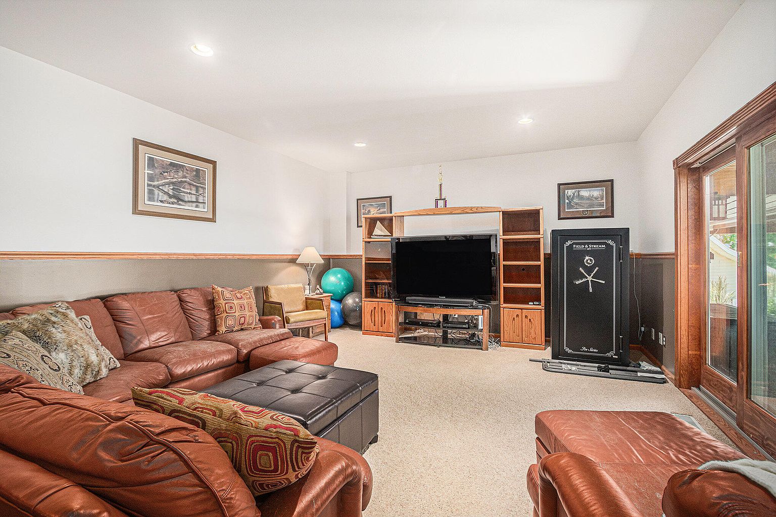 This is a cozy living room featuring a large, brown leather sectional sofa with decorative pillows, a black leather ottoman, and a wooden entertainment center with a large television. A gun safe stands against the wall, and a sliding glass door provides natural light. The room has a warm and inviting atmosphere, perfect for relaxation and entertainment.
