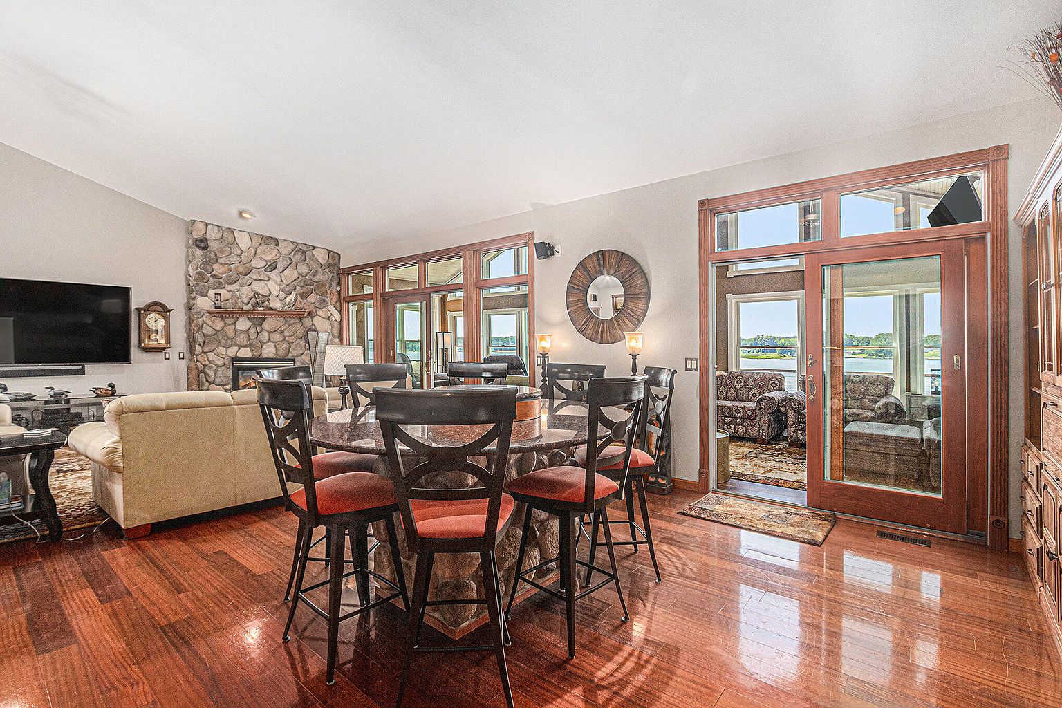 This interior shot showcases a dining area with a round table and chairs, featuring a stone base and dark wood accents. The room boasts hardwood floors and natural light streaming through large windows, offering a glimpse of the outdoors. A stone fireplace and comfortable seating area are visible in the background, adding to the home's inviting atmosphere.