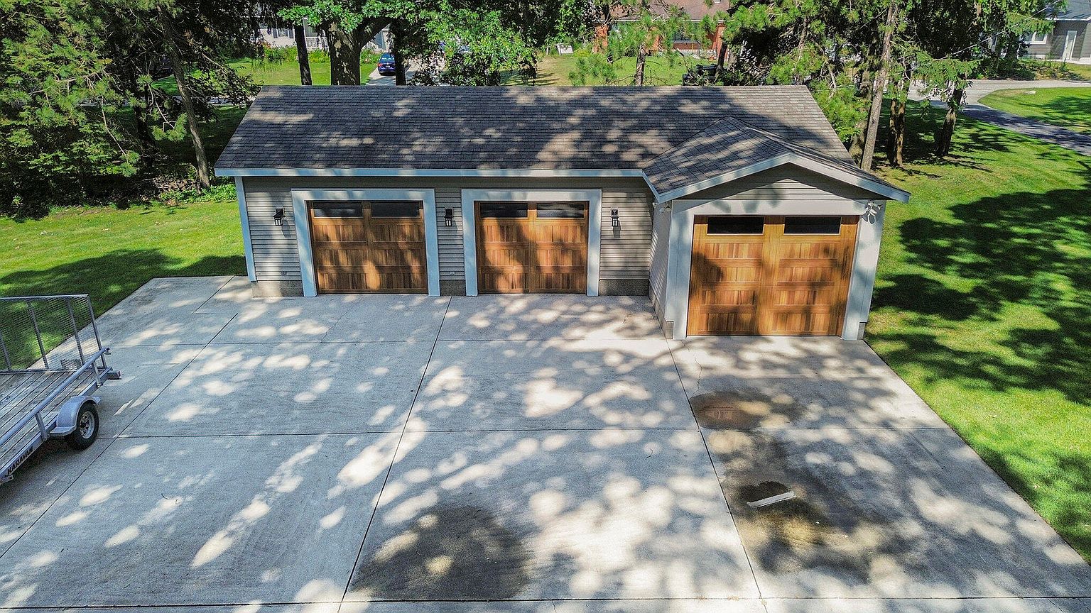 This image showcases a well-maintained detached garage with three wooden doors, set against a backdrop of lush greenery. The concrete driveway is spacious and clean, providing ample parking space. The garage's design complements the surrounding landscape, suggesting a property that is both functional and aesthetically pleasing.