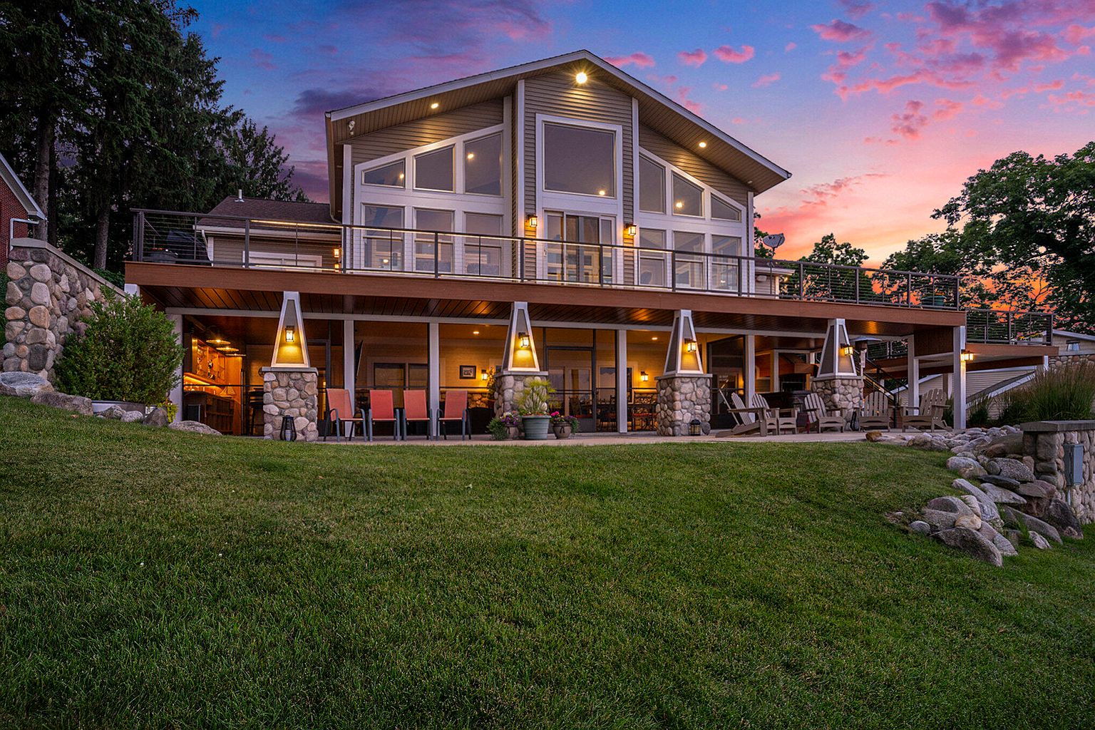 This is a rear exterior view of a two-story house with a large deck. The deck is supported by stone-clad pillars with lights and features a metal railing. The house has large windows and a gable roof, and the lawn slopes gently towards the house.