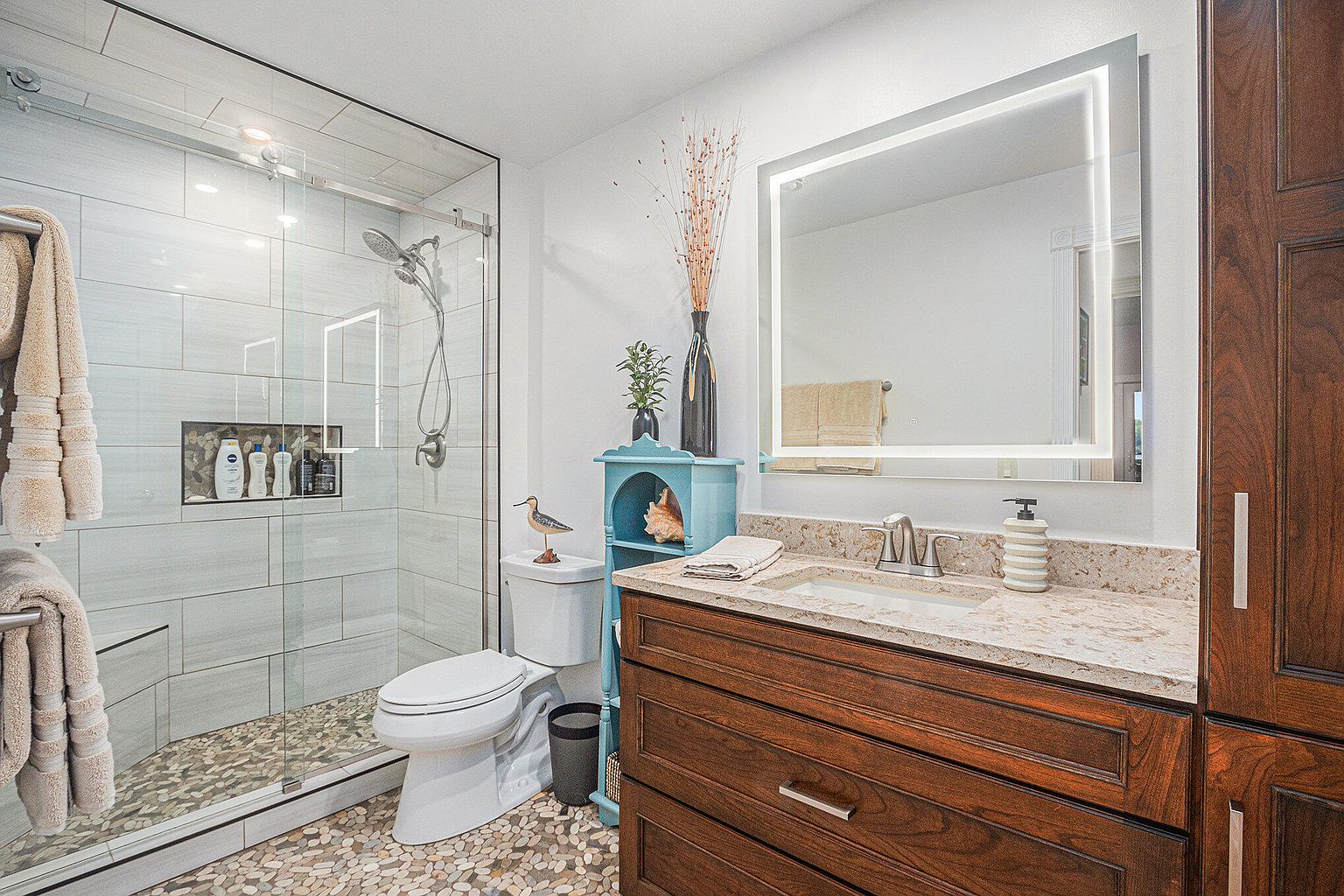 This is a well-lit bathroom featuring a glass-enclosed shower with light gray tiling and a pebble floor. The vanity has a dark wood cabinet with a light-colored countertop, complemented by a square, illuminated mirror. A toilet sits next to a small, turquoise shelving unit, adding a touch of color to the space.