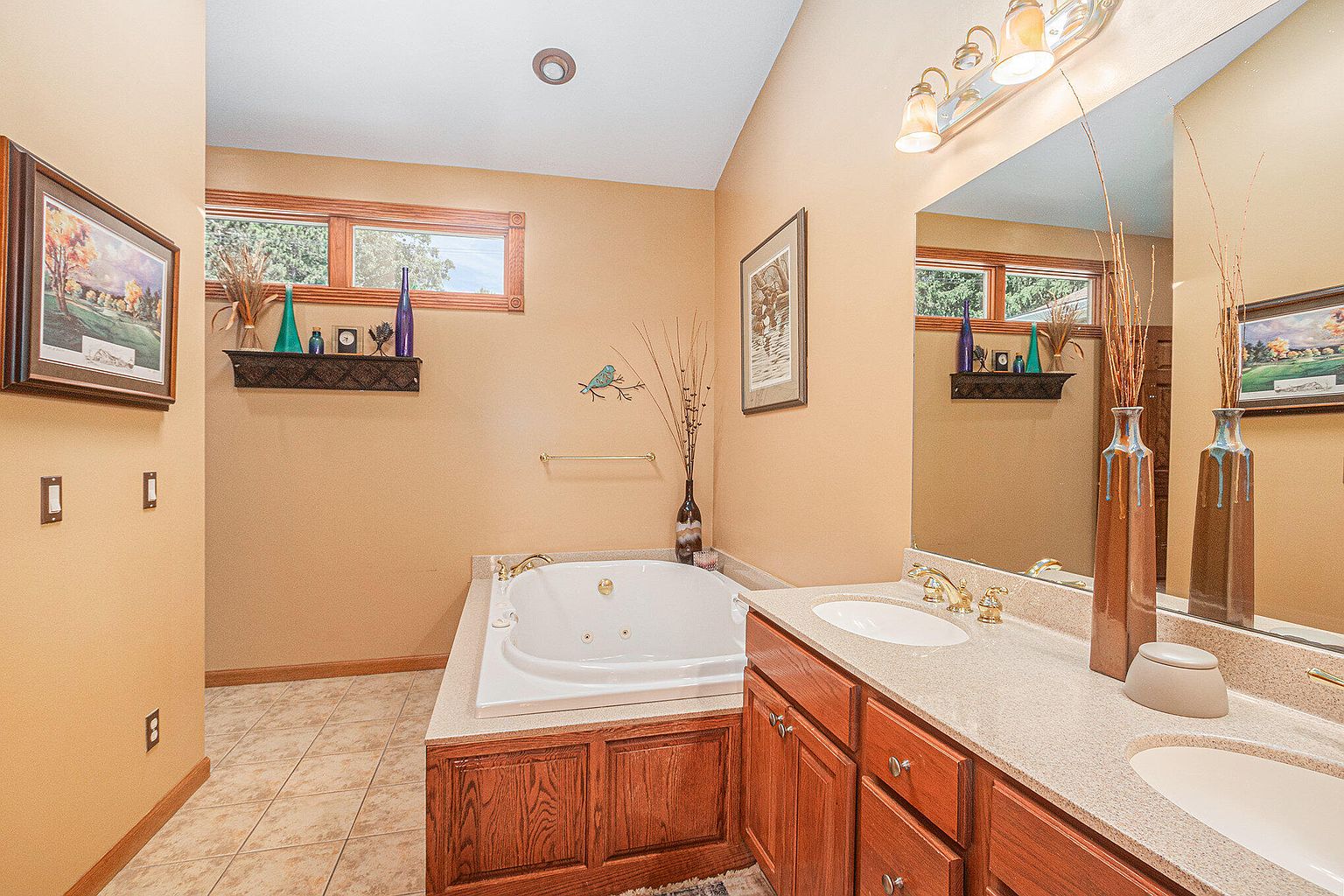 This is a well-lit primary bathroom featuring a jacuzzi tub with wood paneling, a double vanity with light countertops and wooden cabinets, and a large mirror reflecting the window. The walls are painted in a warm tone, and the overall style is traditional and comfortable, suggesting a relaxing and functional space.
