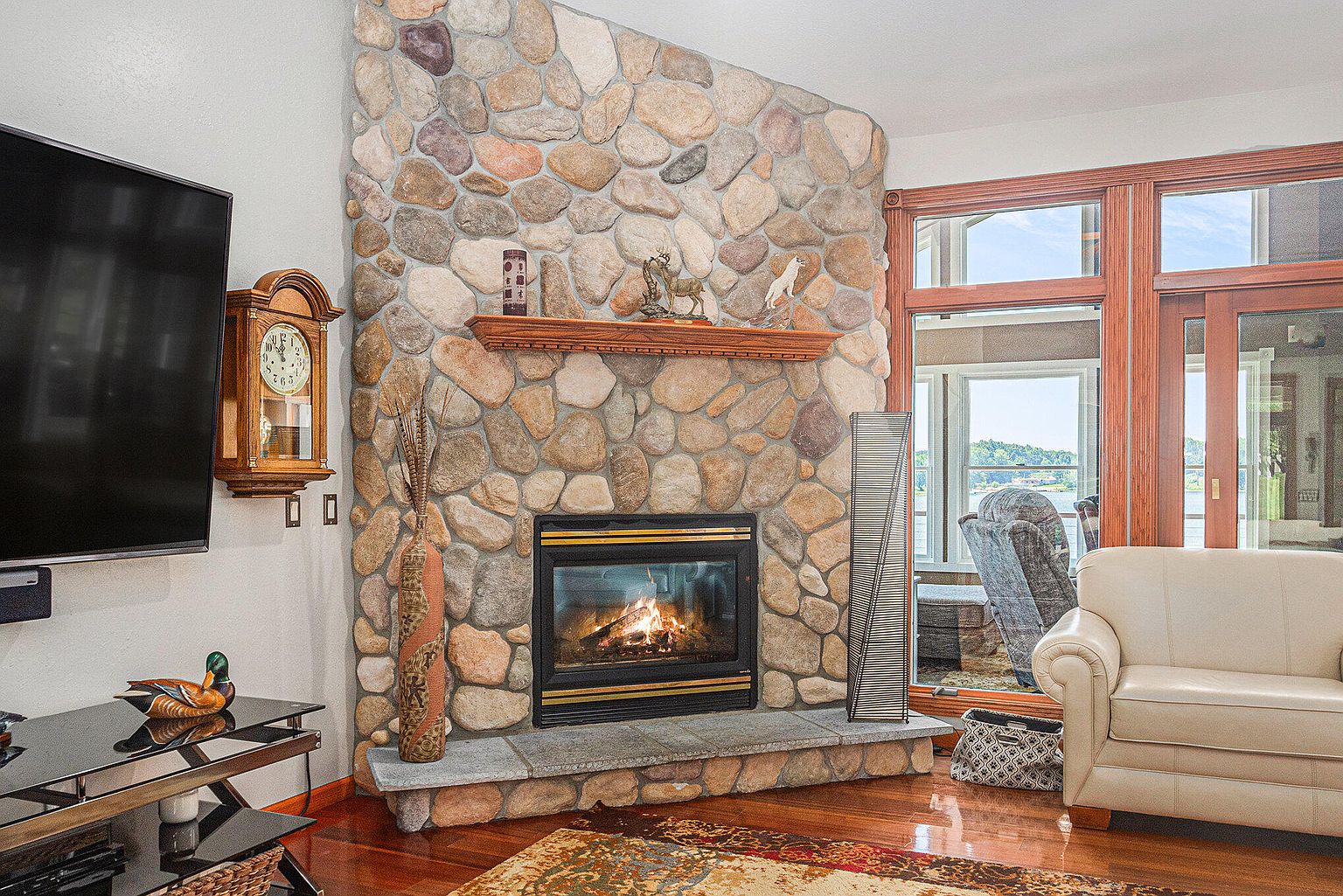 This living room features a stone fireplace as a focal point, complemented by a wooden mantel and hearth. A large television is mounted on the adjacent wall, and a grandfather clock adds a touch of traditional charm. The room is well-lit with natural light streaming through the windows, creating a warm and inviting atmosphere.