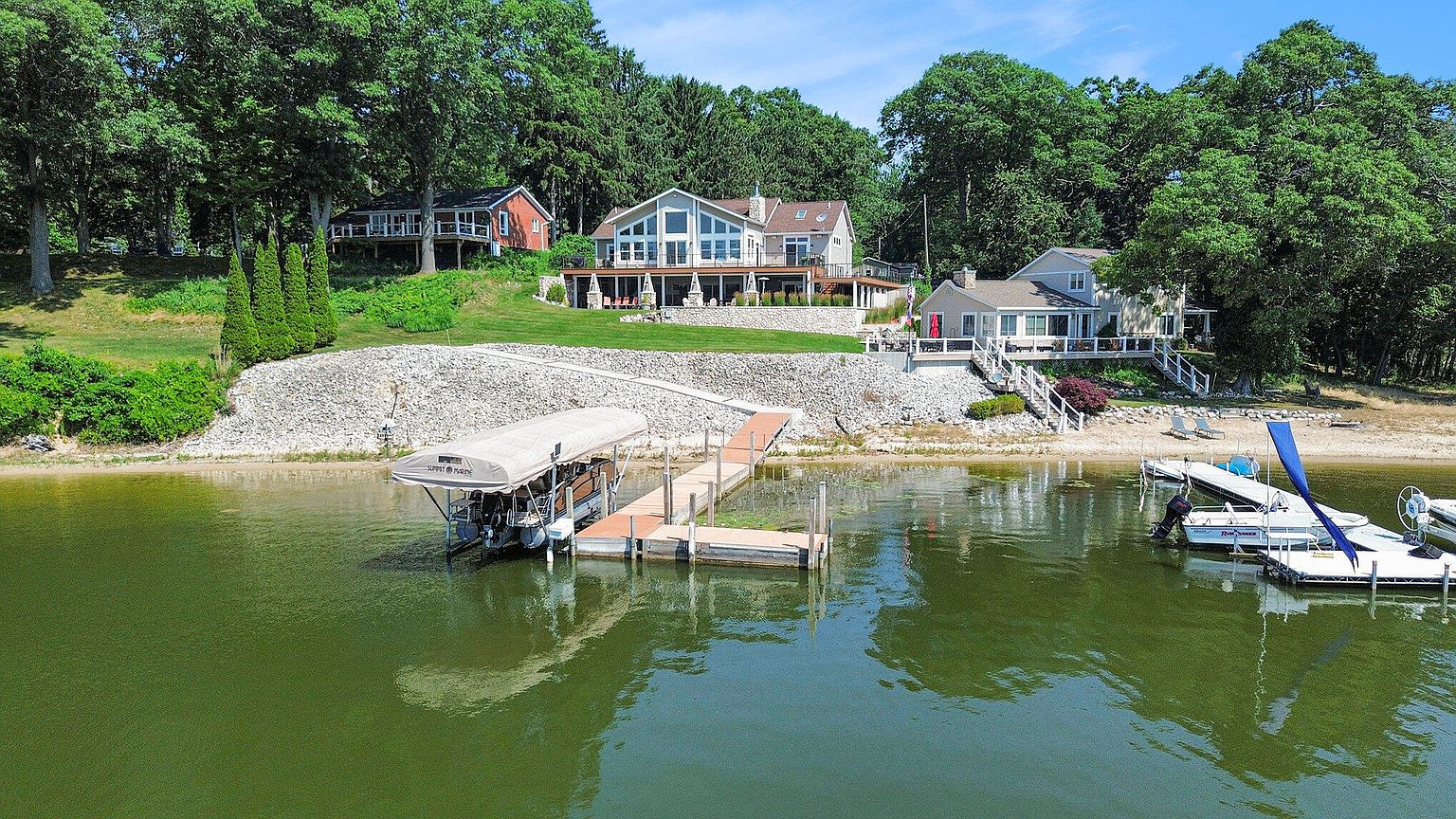 This aerial view showcases a waterfront property with multiple houses nestled among lush trees. A dock extends into the water, featuring a boat lift and several boats. The landscape includes a rocky shoreline and well-maintained lawns, creating a serene and luxurious lakeside setting.