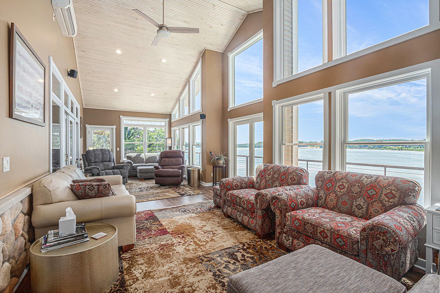 This is a spacious living room with high ceilings and large windows offering a view of the water. The room features a mix of seating options, including sofas and armchairs, arranged on a patterned rug. The decor is comfortable and inviting, creating a relaxing atmosphere with natural light.