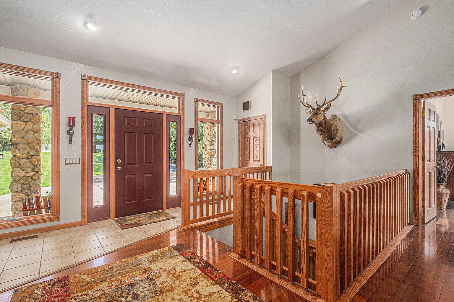 This interior shot showcases a well-lit hallway and staircase area. The space features a combination of tile and hardwood flooring, complemented by a wooden railing and banister. A mounted deer head adds a unique, rustic touch to the decor, while the front door and adjacent windows provide natural light.