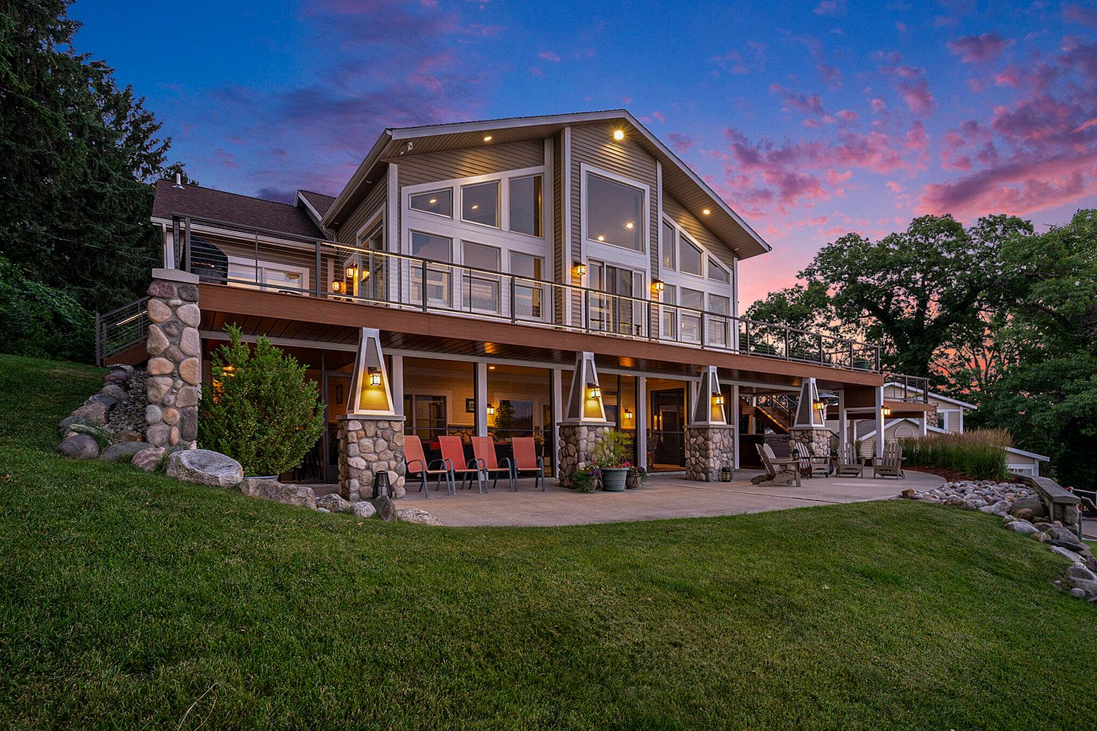 This is a rear view of a two-story house with a large deck. The deck features stone pillars with lights and a metal railing. The house has large windows offering views from the inside, and the yard is well-maintained with green grass and landscaping.