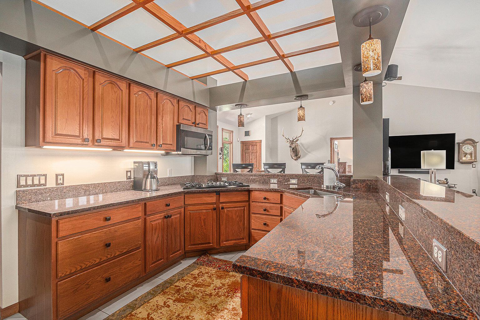 This is a warm and inviting kitchen featuring wooden cabinetry, granite countertops, and a unique coffered ceiling with wood beams. Stainless steel appliances are integrated into the design, and the space opens to a living area with a mounted deer head and television. The overall impression is a blend of rustic charm and modern convenience.