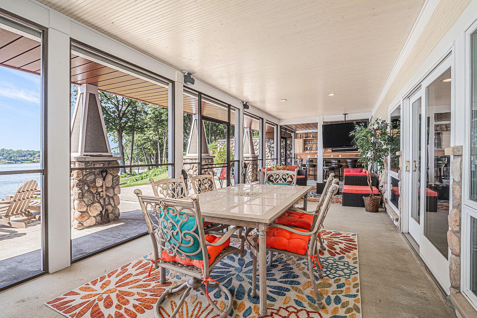 This image showcases a beautifully designed outdoor patio area, featuring a dining set with cushioned chairs and a decorative rug. The space is enclosed with large windows, offering views of the surrounding landscape and water. The patio also includes comfortable seating areas and a bar, creating an inviting and luxurious outdoor living space.
