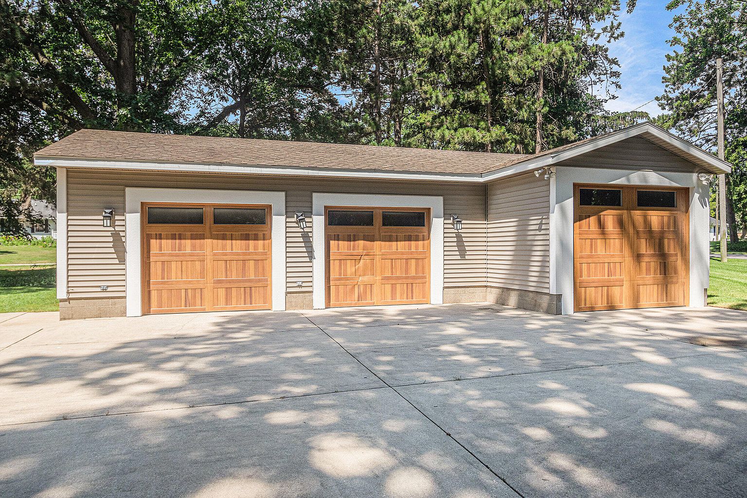 This image showcases a well-maintained three-car garage with wooden doors and light-colored siding. The garage features a concrete driveway and is surrounded by mature trees, suggesting a private and established property. The overall impression is one of quality construction and ample storage space.