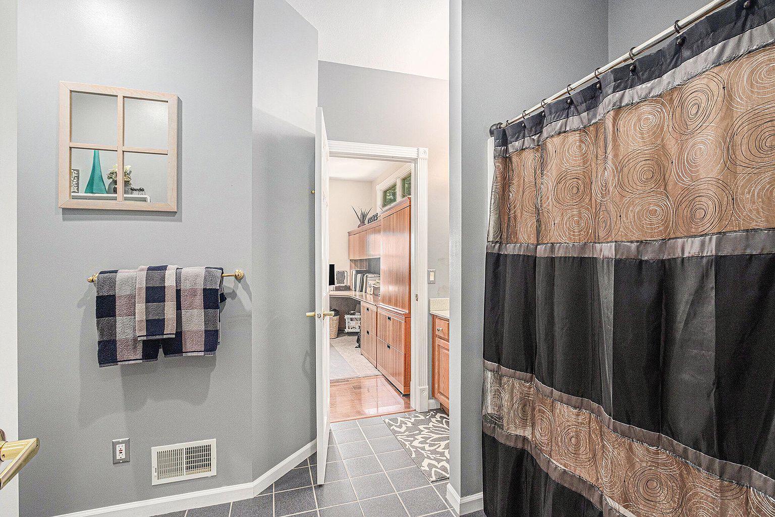 This is a view of a bathroom with gray walls and dark tile flooring. A shower curtain with a circular pattern and black accents is visible on the right. A towel rack with neatly folded towels and a decorative mirror hang on the wall to the left, adding a touch of style to the space.