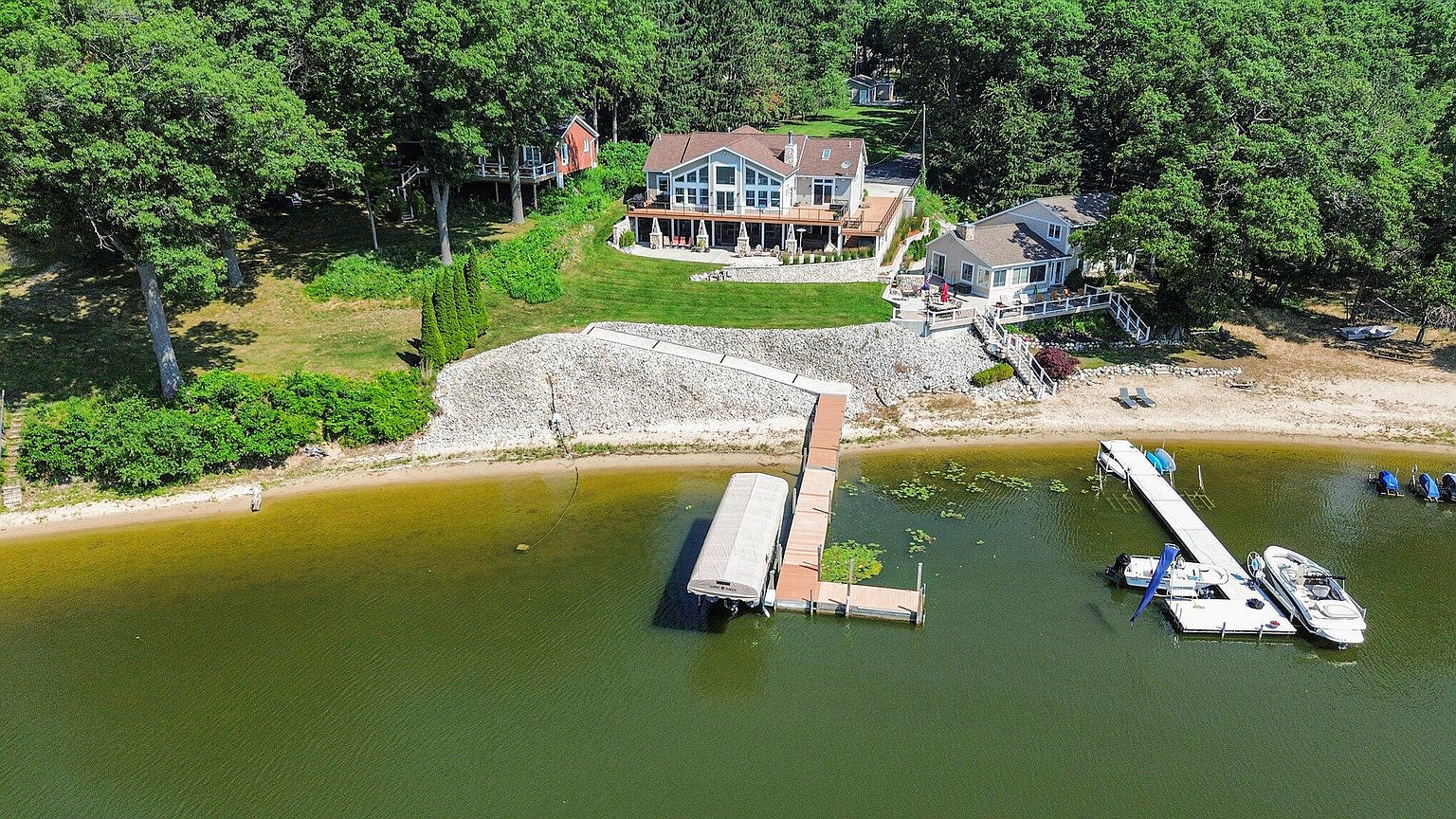 This aerial shot showcases a beautiful lakeside property featuring multiple homes nestled among lush greenery. A large, modern house with a spacious deck overlooks the water, complemented by a smaller house to the side and a charming red house further back. The property boasts a private beach area with docks and boats, creating an idyllic waterfront setting.