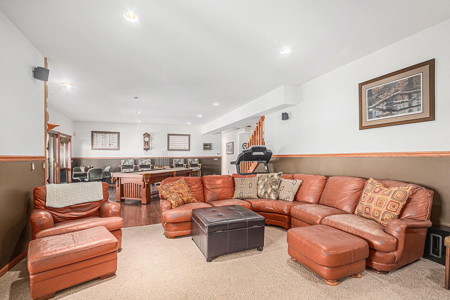 This is a spacious basement living area featuring a large, comfortable leather sectional sofa with decorative pillows, a dark leather ottoman, and a matching leather armchair. The room also includes a pool table, a treadmill, and various wall decorations, creating a recreational and relaxing atmosphere. The flooring is carpeted, and the walls have a two-tone design with a darker lower section.