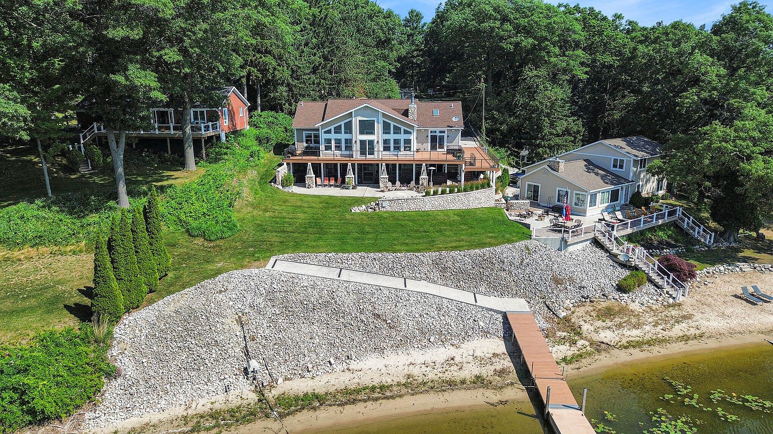 This aerial shot showcases a beautiful lakeside property featuring multiple buildings, including a main house with a large deck, a smaller cottage, and another house to the side. The property boasts a well-maintained lawn, stone landscaping, and a private dock extending into the lake. The surrounding mature trees provide privacy and a serene atmosphere, making it an ideal waterfront retreat.