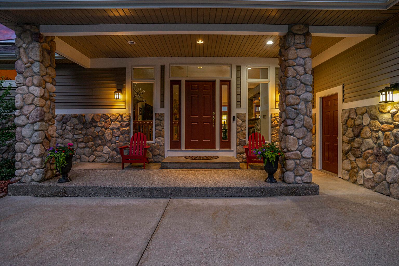 This is a well-lit entryway of a home, featuring a covered porch with stone pillars and a red front door. Two red Adirondack chairs flank the entrance, adding a touch of color and inviting appeal. The stone accents and warm lighting create a welcoming and sophisticated first impression.