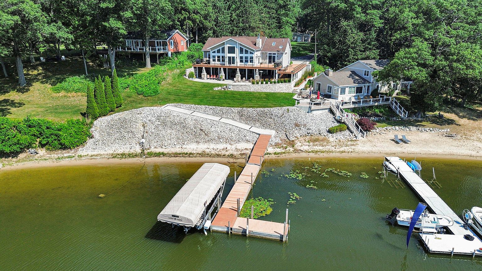 This aerial shot showcases a beautiful lakeside property featuring multiple buildings, including a main house with a large deck, a smaller cottage, and a boathouse with a covered boat slip. The property boasts a well-maintained lawn, a stone retaining wall leading to the waterfront, and docks for boating and water activities. The surrounding mature trees provide privacy and a serene backdrop, enhancing the property's appeal as a luxurious waterfront retreat.