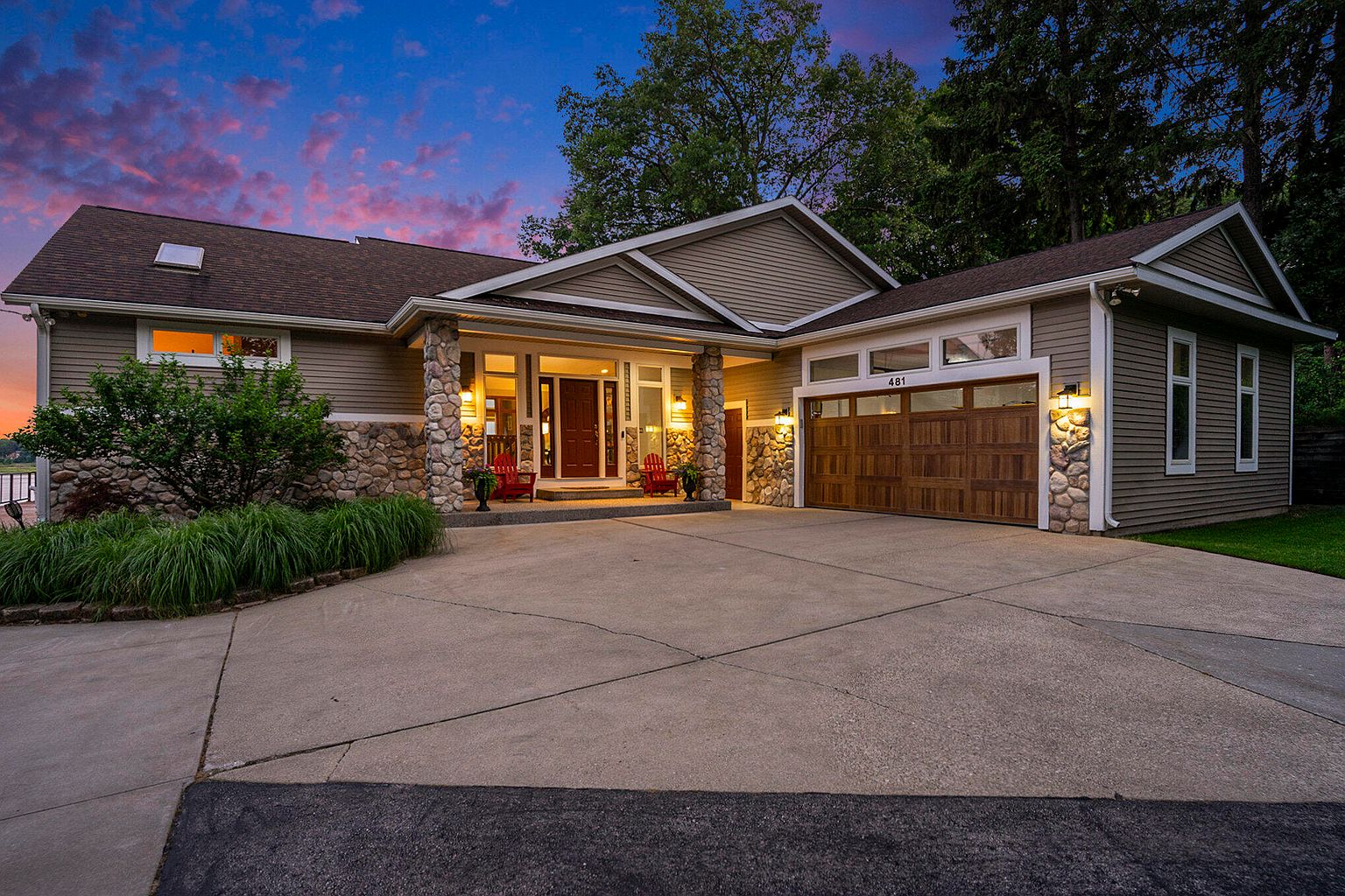 This is a front exterior view of a well-maintained single-family home. The house features a combination of stone and siding, a two-car garage with wooden doors, and a covered front porch with red chairs. The driveway is spacious, and the landscaping includes neatly trimmed bushes and trees, creating a welcoming and upscale impression.