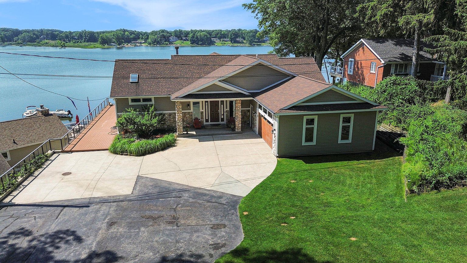 This aerial view showcases a charming single-family home with a well-manicured lawn and a spacious driveway. The house features a combination of stone and siding exterior, a brown shingle roof, and an attached garage. A wooden deck extends towards the waterfront, offering stunning views of the lake and surrounding greenery, enhancing the property's appeal as a lakeside retreat.