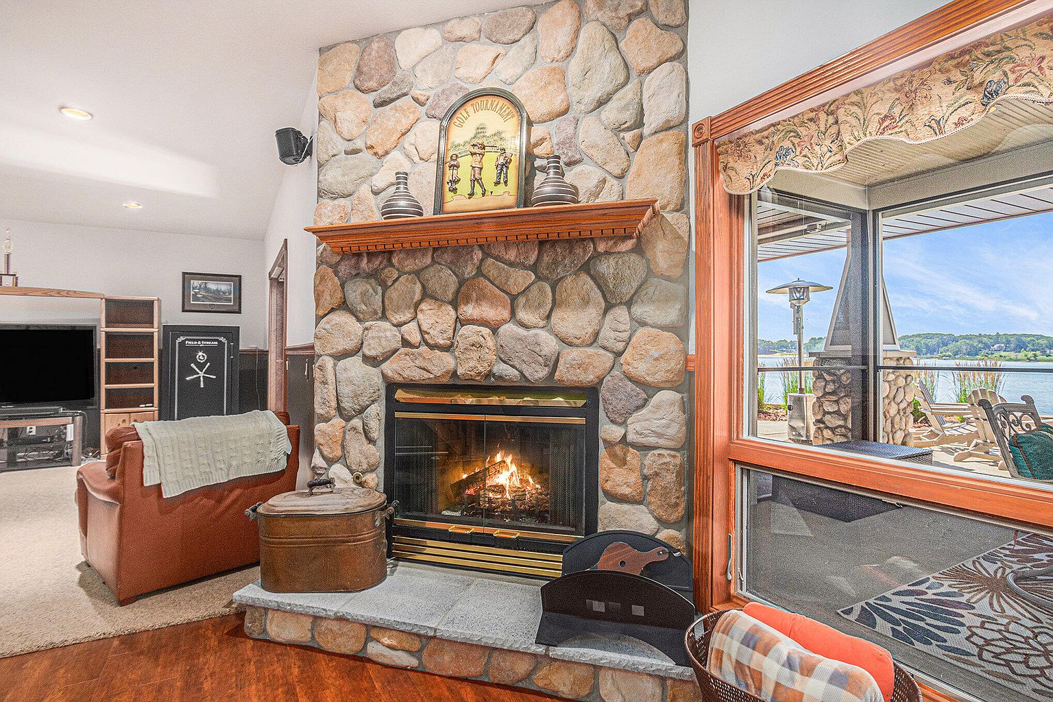 This inviting living room features a stone fireplace as its focal point, complemented by a wooden mantel and decorative items. A comfortable leather armchair sits nearby, and a large window offers a view of the outdoors, enhancing the room's natural light. The overall ambiance is cozy and relaxing, perfect for a family home.