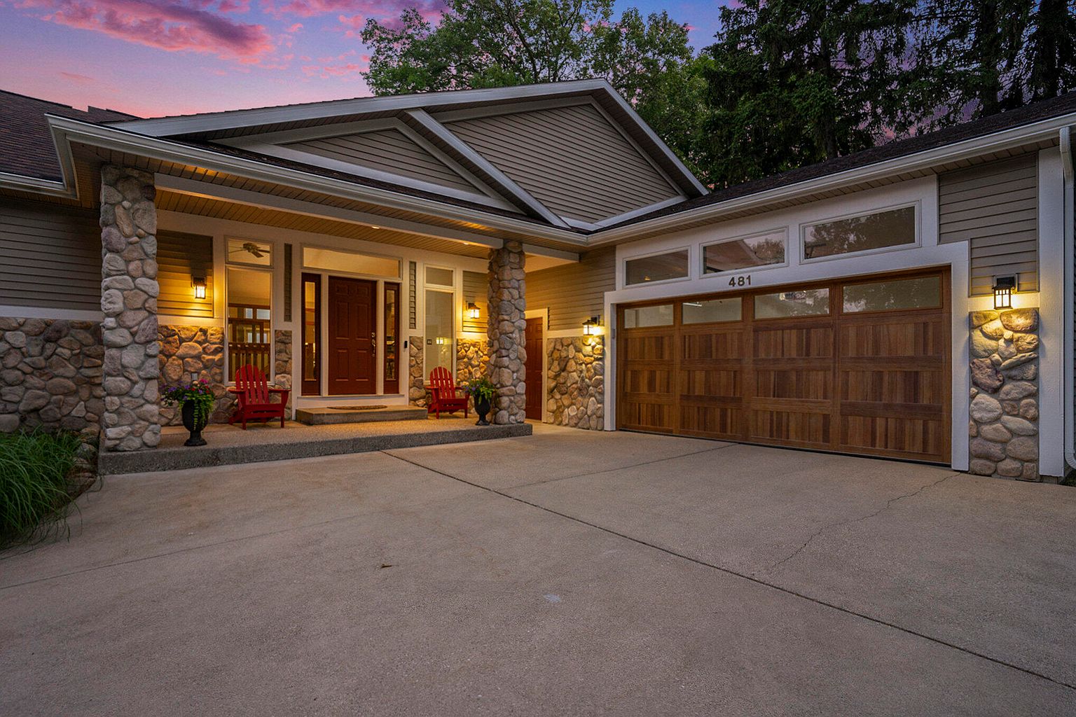 This is a front exterior view of a single-family home featuring a combination of stone and siding. The house has a prominent front porch with stone pillars and two red Adirondack chairs. A two-car garage is visible on the right side of the house, and the driveway leads up to the garage and front entrance.