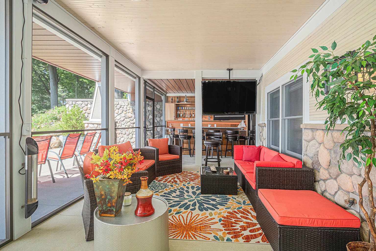 This image showcases a well-appointed outdoor patio area, featuring comfortable wicker furniture with vibrant orange cushions, a stylish area rug, and a built-in bar in the background. The space is partially enclosed with screens, offering a blend of indoor and outdoor living, and includes a mounted television for entertainment. The stone accents on the walls and the view of the surrounding greenery add to the patio's appeal.