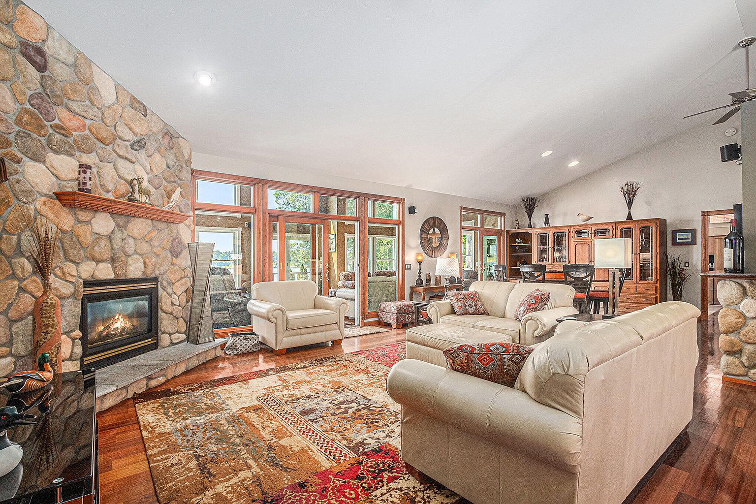 This is an interior shot of a spacious living room featuring a stone fireplace as a focal point. The room is furnished with comfortable, light-colored sofas and a patterned area rug, creating a warm and inviting atmosphere. Large windows provide ample natural light and views of the outdoors, enhancing the room's appeal.