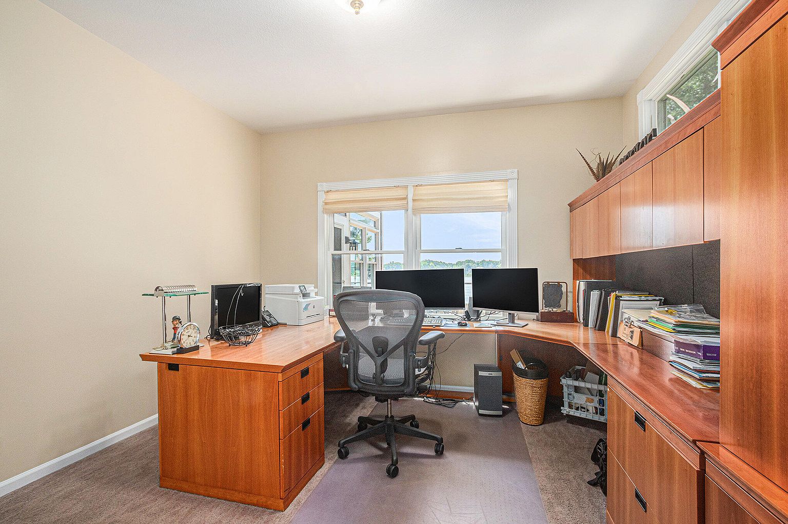This is an interior shot of a home office featuring a large wooden desk with built-in cabinets and drawers. The desk is equipped with dual monitors, a printer, and various office supplies. An ergonomic office chair sits in front of the desk, and natural light streams in through a window, creating a bright and functional workspace.