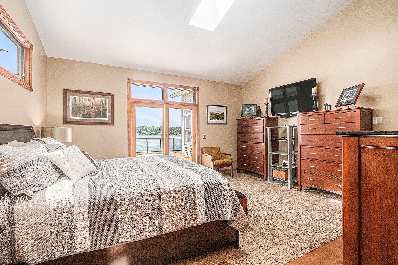 This is a primary bedroom featuring a king-size bed with a patterned comforter and several pillows. The room has a warm, neutral color palette with wood furniture, including dressers and a side table. A large glass door leads to a balcony with a lake view, and a skylight provides natural light.